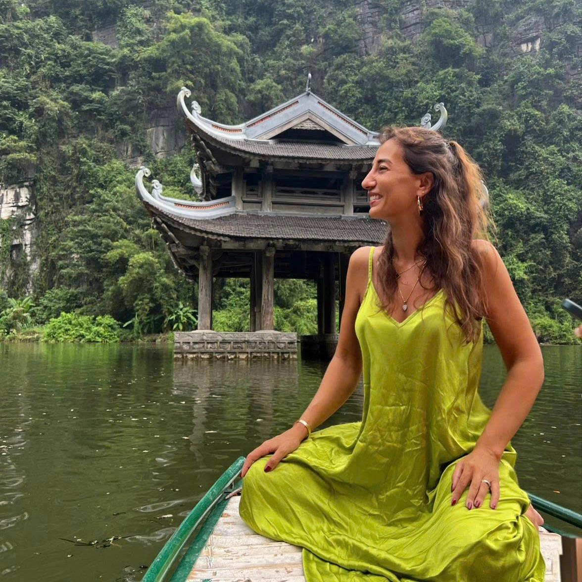 A woman in a yellow dress sitting at the prow of a boat in front of a lakeside pavilion