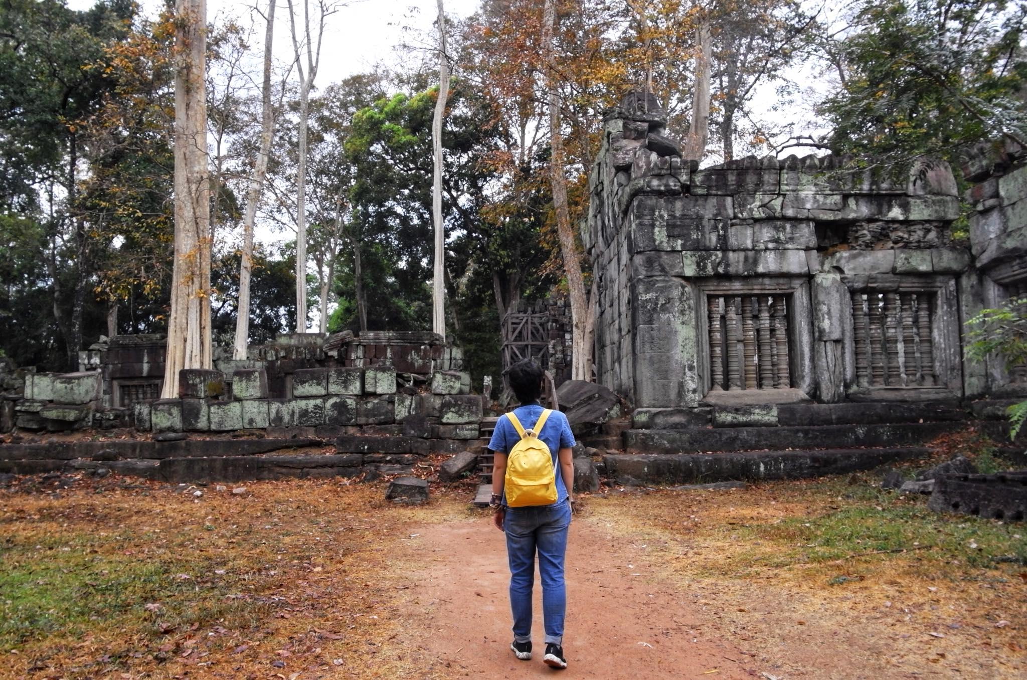 A traveller with a yellow backpack walking a dirt path towards an overgrown temple ruin