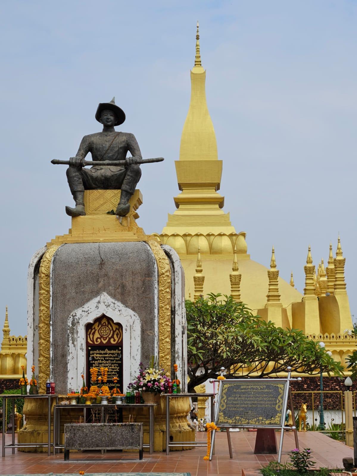 A golden stupa rising behind a royal statue with offerings at its base