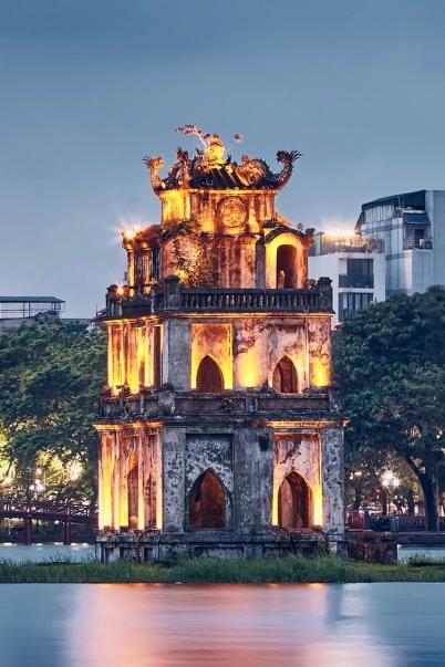 A floodlit stone tower on a small island in a city lake at dusk
