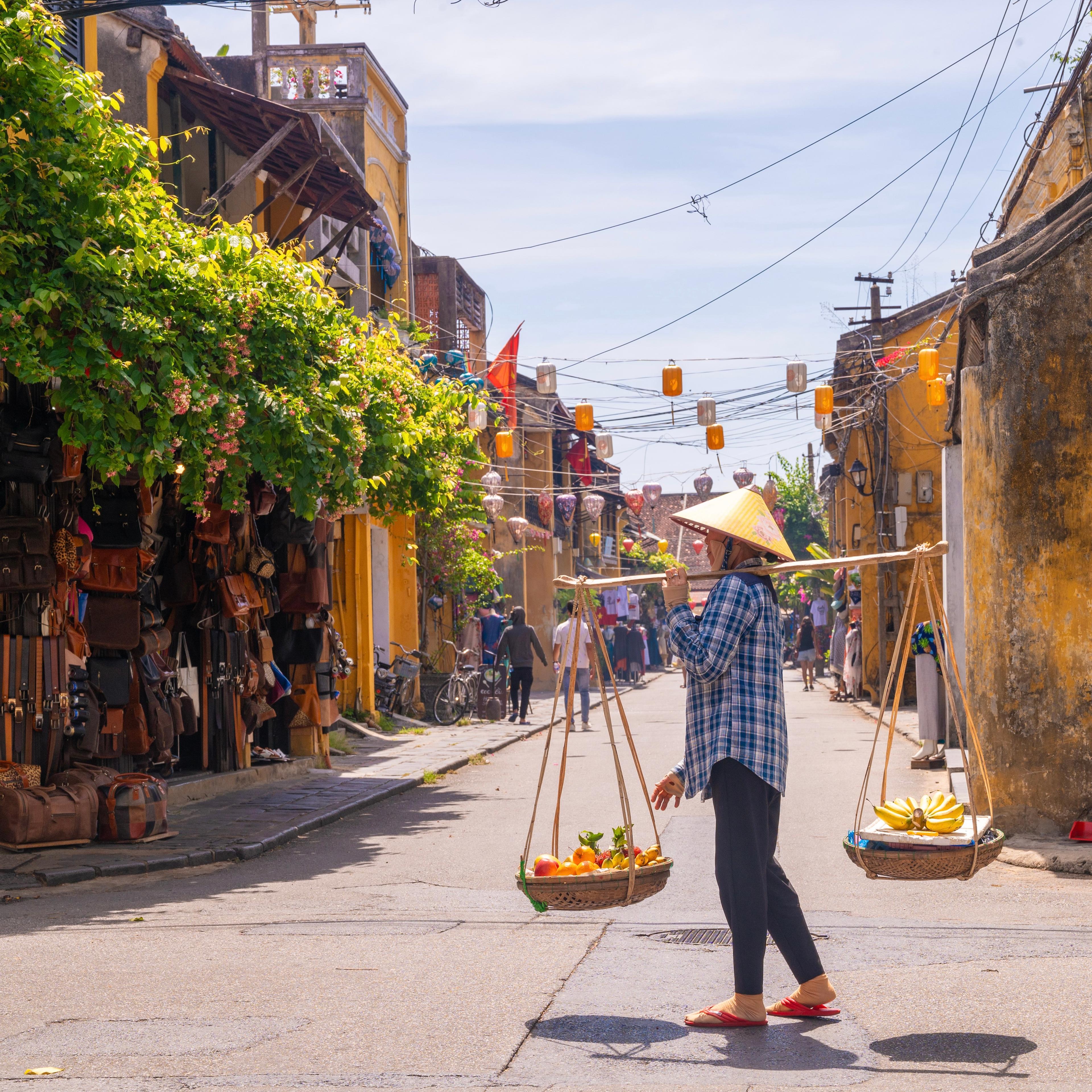 A street vendor carrying baskets on a shoulder pole past lantern-hung shopfronts in an old town street