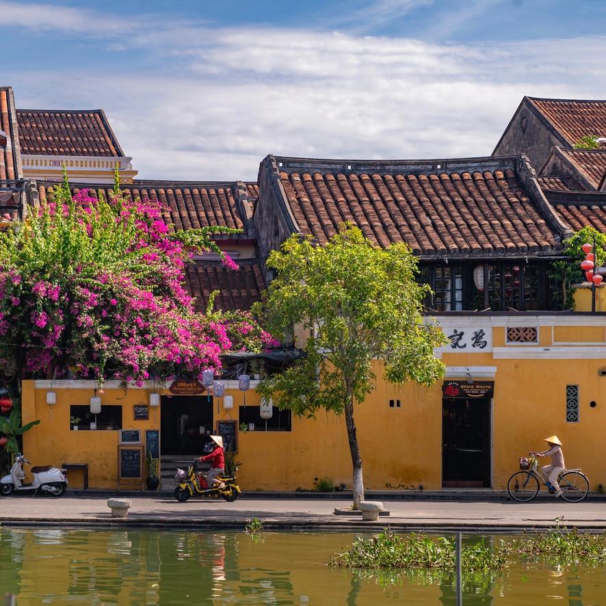 Yellow shophouses with tiled roofs and pink bougainvillea beside a canal, a cyclist in a conical hat riding past