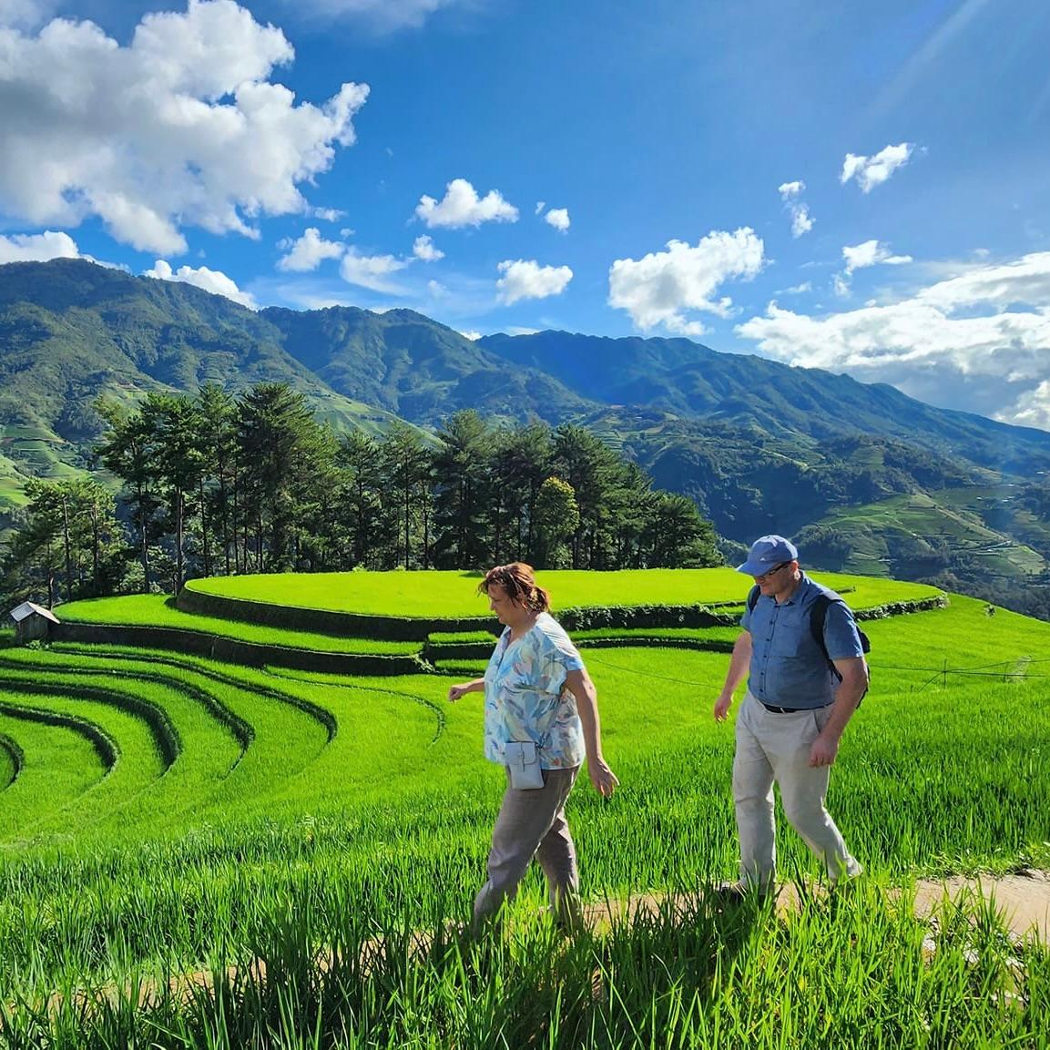 Two travellers walking a path through green terraced rice fields below forested mountains