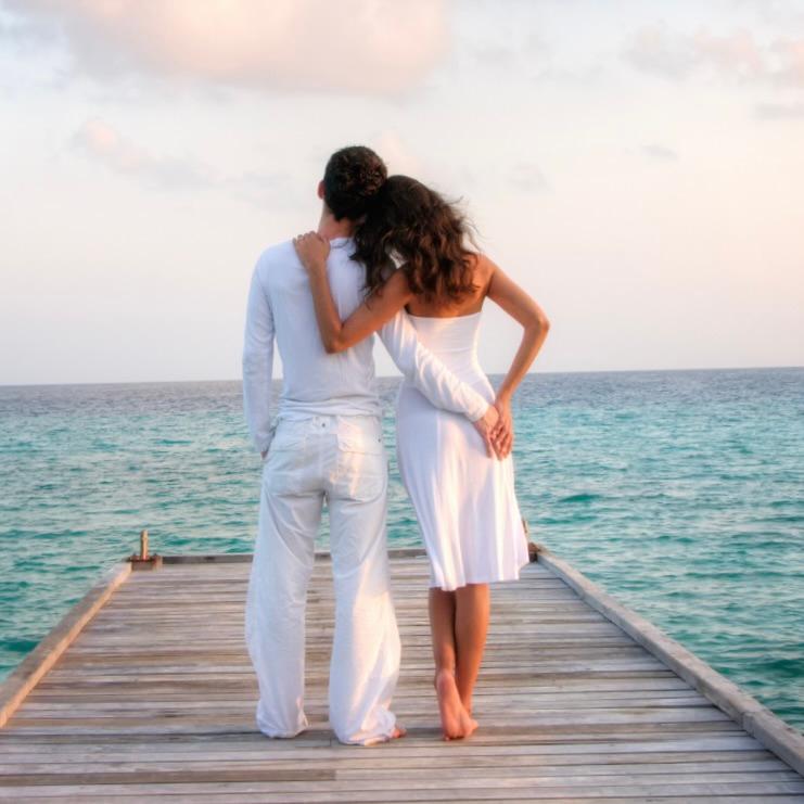 A couple dressed in white standing arm in arm at the end of a wooden jetty above turquoise water