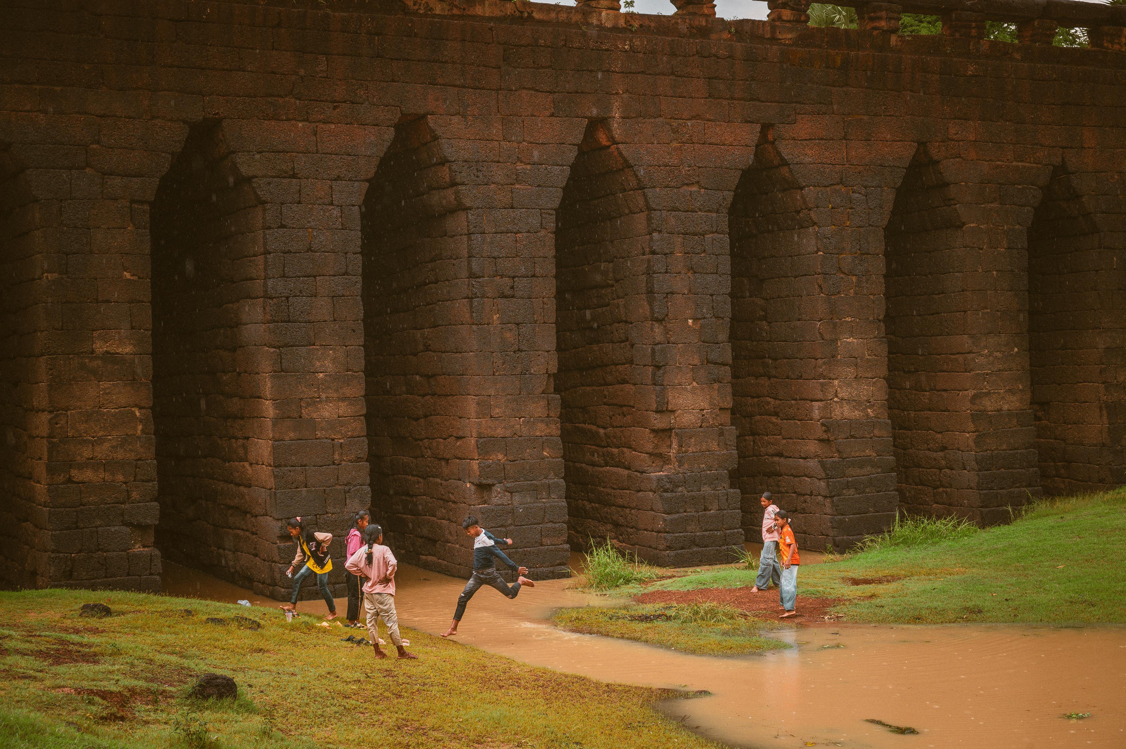 Children running along a sandy bank beside the arches of an old stone bridge