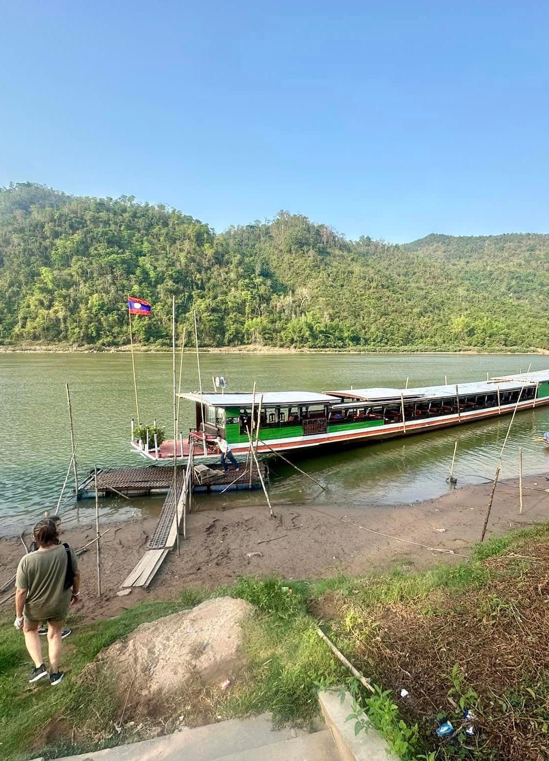 A long river boat moored at a bank below forested hills