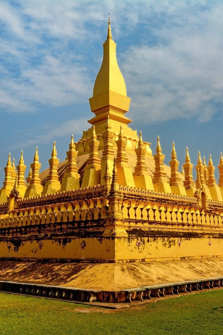 A golden stupa with a tall spire ringed by rows of smaller spires, on a lawn under a blue sky