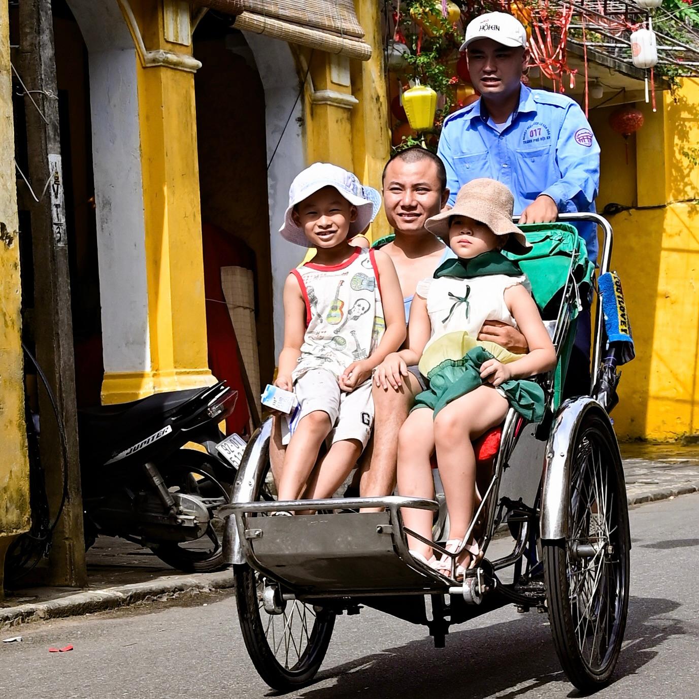 Two children riding with an adult in a cyclo past yellow shophouses