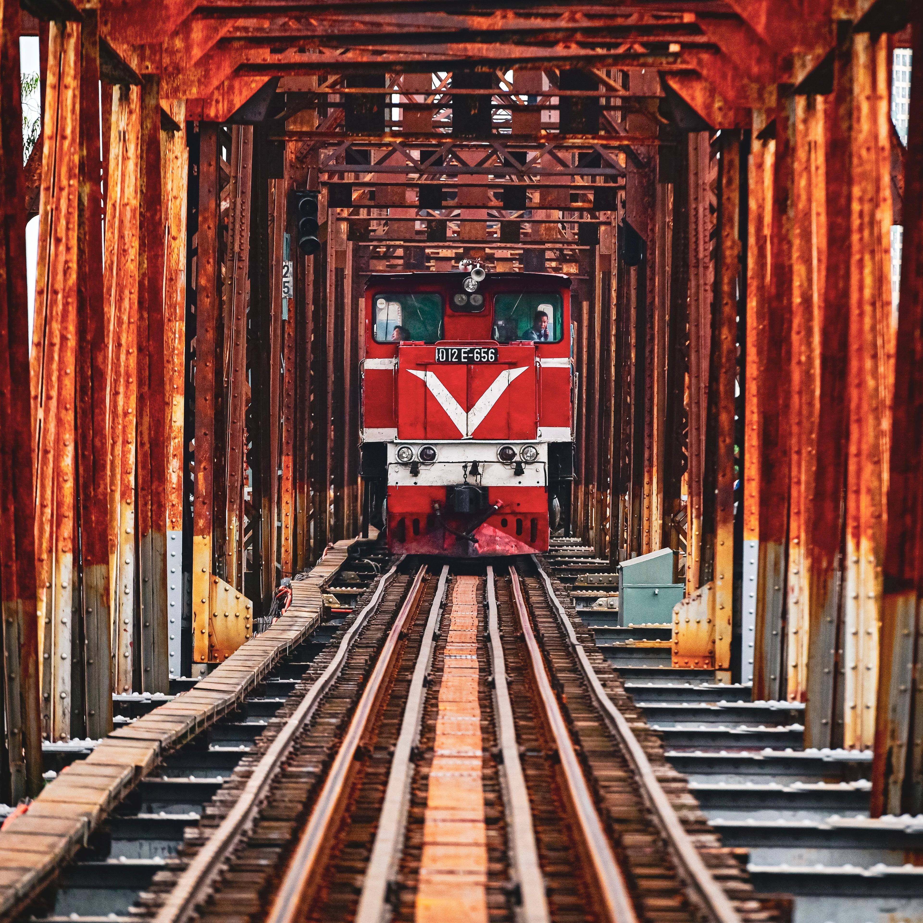A red locomotive crossing an old steel railway bridge, seen head-on between the girders